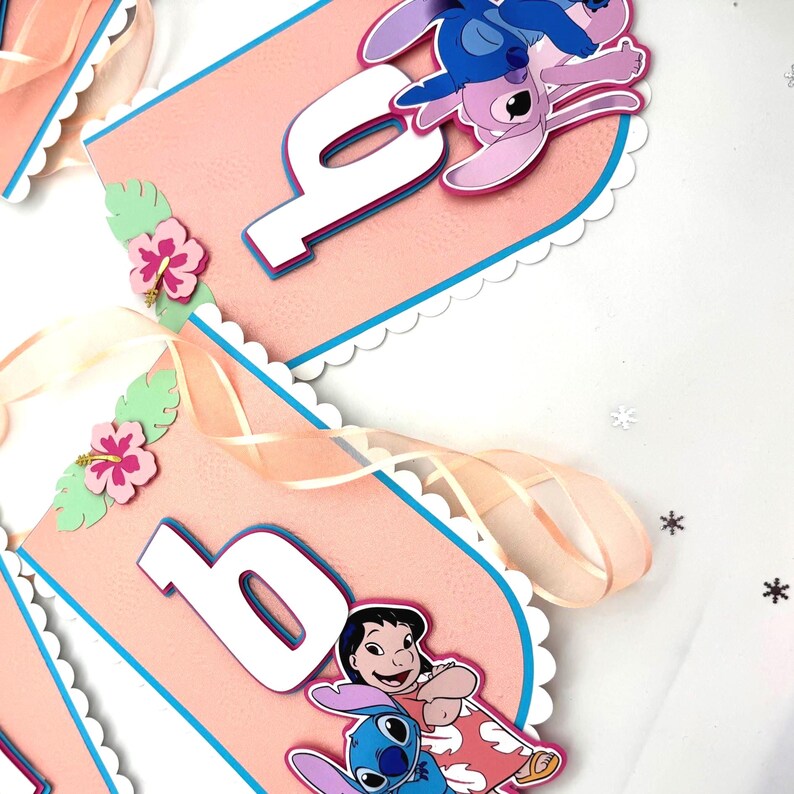 May include: A pink and blue birthday banner with the number '6', white borders, Stitch and Lilo characters, and hibiscus flower decorations. The banner has a scalloped edge and is strung with a sheer ribbon.