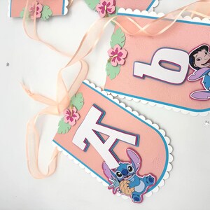 May include: Pink banner with white scalloped edges, blue trim, and white letters. Features a cartoon character and tropical flower accents. A sheer ribbon is attached for hanging. Party decoration.