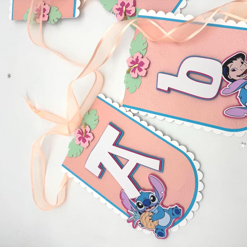 May include: Pink banner with white scalloped edges, blue trim, and white letters. Features a cartoon character and tropical flower accents. A sheer ribbon is attached for hanging. Party decoration.