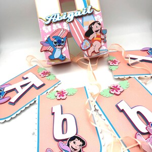 May include: A custom party decoration with a tropical theme. The centerpiece is a white and pink house-shaped box with the name "Abigail". The box is decorated with cartoon characters, surfboards, and a palm tree. A banner spells out "Abigail".