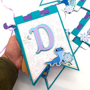 May include: A teal and purple banner with the letter "D" in light purple and a blue lizard character. The banner has a white background with a snowflake pattern and is strung with a purple ribbon.