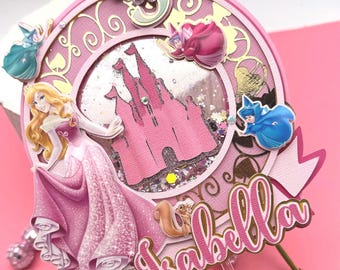 May include: A circular cake topper featuring a pink castle shaker element, a blonde princess in a pink gown, three small fairies in blue, red, and green, and a squirrel. The number 3 is at the top, and the name Isabella is in pink script at the bottom.