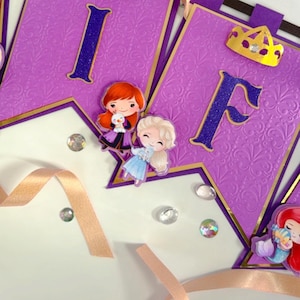 Op de afbeelding: Paarse banner met gouden rand en reliëf bloemmotief. Bevat de letters "I" en "F" in glinsterend blauw. Inclusief cartoonafbeeldingen van prinsessen en een gouden kroon. Decoratief lint en sprankelende edelstenen maken het ontwerp compleet.
