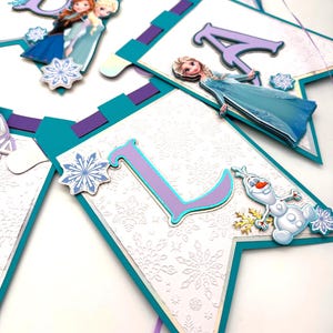 May include: A banner with the letters "A", "L", and "O" in purple and teal, featuring images of Elsa, Anna, and Olaf from the movie Frozen. The banner has a white background with a snowflake pattern and teal and purple accents.