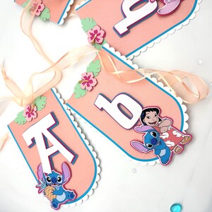 May include: A pink banner with white scalloped edges and blue trim. The banner features the letter "A" and the number "6" in white, along with cartoon characters and floral accents. A sheer ribbon is attached for hanging.