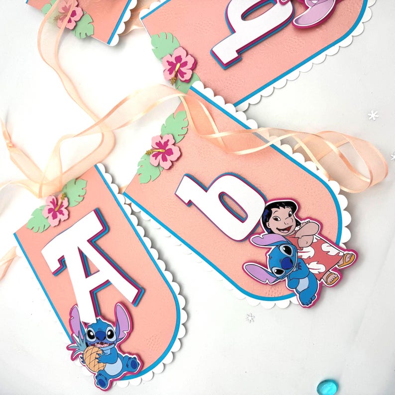 May include: A pink banner with white scalloped edges and blue trim. The banner features the letter "A" and the number "6" in white, along with cartoon characters and floral accents. A sheer ribbon is attached for hanging.
