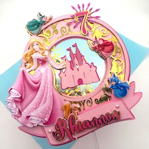 May include: A circular cake topper featuring a pink princess in a gown, a pink castle silhouette, and three small fairies. The design includes a glittery number 7, gold filigree accents, and a pink banner with the name Rhiannon in script.
