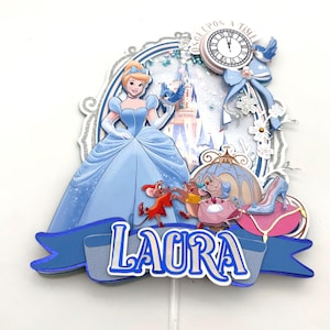 May include: A light blue Cinderella-themed cake topper with the name "LAURA" in white and blue. The design includes Cinderella, a clock, a carriage, mice, and a glass slipper. The words "ONCE UPON A TIME" are also visible.