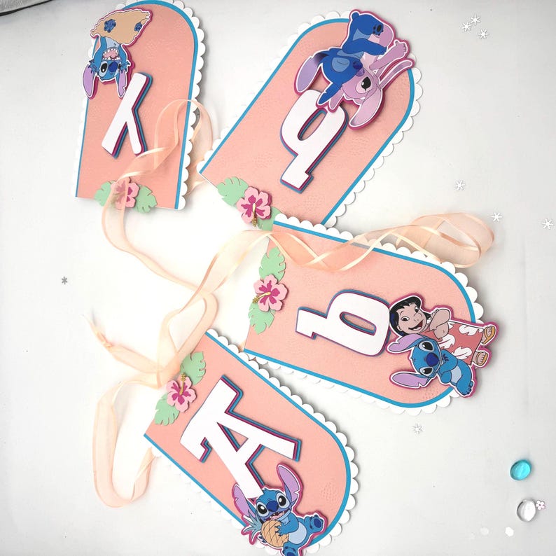 May include: Pink banner with white scalloped edges and blue trim, featuring the letters "K", "A", and "Q". Each banner has a Stitch character illustration and floral accents. Light pink ribbon is attached for hanging.