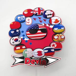 May include: A colorful cake topper featuring countryball designs representing various nations. The central countryball is the United States, wearing sunglasses. The number "9" is displayed, and the name "Devin" is written on an arrow-shaped base.