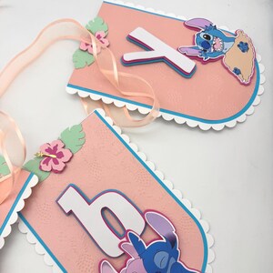 May include: Pink banner with white scalloped edges, blue trim, and a light pink ribbon. Features the letter "Y" and a cartoon character. Adorned with pink flowers and green leaves. The banner is likely for a birthday or party.