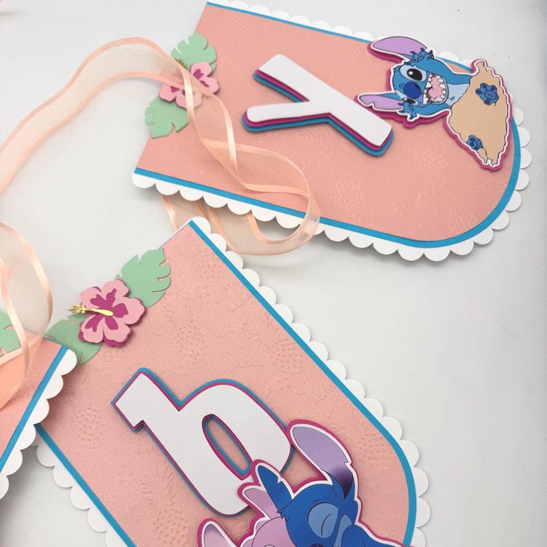 May include: Pink banner with white scalloped edges, blue trim, and a light pink ribbon. Features the letter "Y" and a cartoon character. Adorned with pink flowers and green leaves. The banner is likely for a birthday or party.