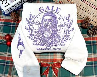 Op de afbeelding: Witte trui met een paars geborduurd ontwerp met een personage genaamd Gale uit Baldur's Gate. Het ontwerp bevat een krans, sterren en wolken.