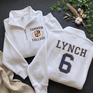 May include: Two white quarter-zip sweatshirts. The left sweatshirt features an embroidered crest with the text "TOMMEN COLLEGE". The right sweatshirt displays the name "LYNCH" in large navy letters above the number "6" in navy and gold.