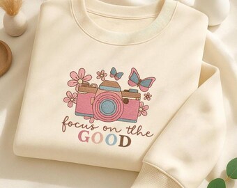 May include: Cream-colored sweatshirt with a retro camera graphic in pink, blue, and brown, surrounded by flowers and butterflies. The text "focus on the GOOD" is printed below the camera. The sweatshirt has a ribbed collar and cuffs.
