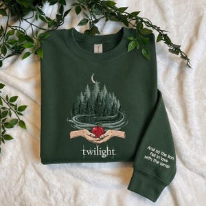 Puede incluir: Una sudadera de cuello redondo verde oscuro con un gráfico de dos manos sosteniendo una manzana roja bajo un bosque y una luna creciente. La palabra twilight está impresa debajo de las manos y la manga lleva el texto And so the lion fell in love with the lamb.