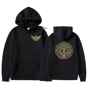 Puede incluir: Sudadera con capucha negra con un emblema dorado y un diseño circular dorado. La sudadera tiene un bolsillo delantero y una capucha con cordón. El diseño está inspirado en un videojuego popular. Medidas en centímetros.