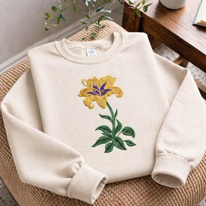 以下が含まれることがあります： クリーム色のスウェットシャツ。黄色いユリの花と紫色のアクセントが刺繍されています。花には緑色の茎と葉があります。スウェットシャツは織りの椅子に展示されています。