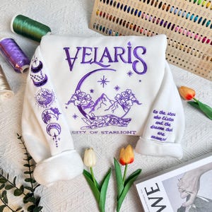 Può includere: Felpa bianca con ricamo viola che rappresenta una luna crescente, montagne e il testo "VELARIS CITY OF STARLIGHT". La manica presenta un ricamo viola aggiuntivo di lune e stelle.