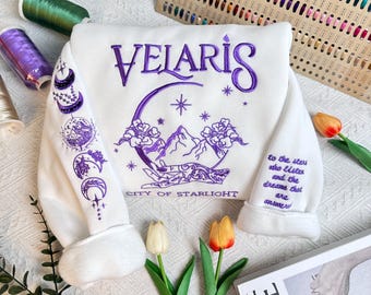 May include: White sweatshirt with purple embroidery featuring a crescent moon, mountains, and the text "VELARIS CITY OF STARLIGHT". The sleeve has additional purple embroidery of moons and stars.