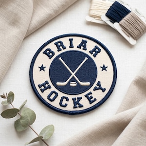 May include: A circular embroidered patch with the text "BRIAR HOCKEY" in navy blue. The patch features a navy blue circle with crossed hockey sticks and a puck. It is bordered by a navy blue ring and stars, suitable for clothing.