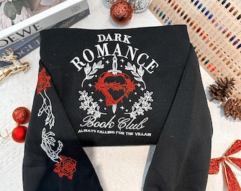 May include: Black sweatshirt with white embroidery that reads "Dark Romance Book Club Always Falling for the Villain". The design features a red heart with a dagger through it and a crown of thorns.