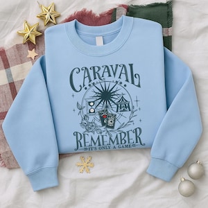 May include: A pale blue sweatshirt featuring the text "CARAVAL REMEMBER IT'S ONLY A GAME". The design includes a Ferris wheel, playing cards, and floral motifs. Gold star and snowflake decorations are in the background.