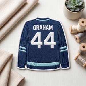 Könnte beinhalten: Ein marineblaues Hockey-Trikot-Patch mit dem Namen "GRAHAM" und der Nummer "44" in Weiß. Das Trikot hat hellblaue Streifen an den Ärmeln und am Kragen. Der Patch ist mit einem weißen Rand bestickt.