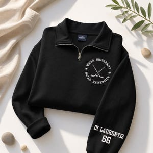 May include: Black quarter-zip sweatshirt with the text "Briar University" and crossed hockey sticks in a circular design. The sleeve features "Di Laurentis 66" in white. The sweatshirt has a collared neckline and a silver zipper.