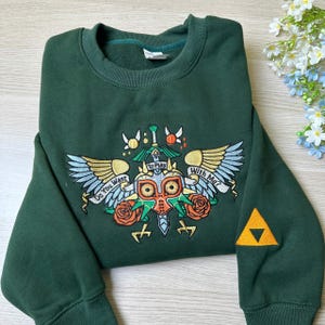 May include: Dark green sweatshirt with an embroidered design featuring a mask, wings, roses, and the text "Do You Want To Play With Me?". A gold triangle is on the sleeve.