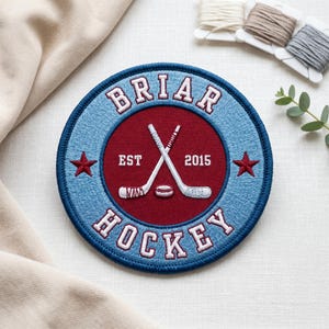May include: A circular embroidered patch with the words "Briar Hockey" in white lettering on a blue and red background. The patch features crossed hockey sticks and a puck, with the year "2015" and two red stars.