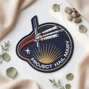 May include: Embroidered patch with a navy blue background featuring a stylized spaceship design. The patch includes the text "PROJECT HAIL MARY" in white lettering, with a gold sunburst design at the bottom and a star.