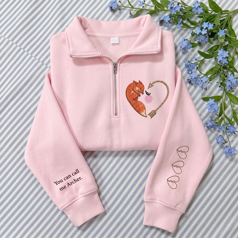 May include: A light pink quarter-zip sweatshirt featuring an embroidered orange fox curled into a heart shape with an arrow and a small pink apple. The right sleeve displays the text "You can call me Archer." and the left sleeve has three hearts.