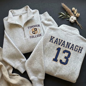 Puede incluir: Dos sudaderas grises claras con cuello con cremallera. Una muestra "TOMMEN COLLEGE" con un escudo, la otra tiene "KAVANAGH 13" en azul marino. Las sudaderas están sobre una superficie oscura con elementos decorativos.