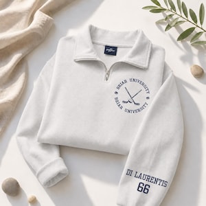 May include: A light gray sweatshirt with a quarter-zip collar. The sweatshirt features a navy blue circular emblem with crossed hockey sticks and the words "Briar University." The sleeve displays "Di Laurentis 66" in navy blue. The sweatshirt is folded on a white surface.