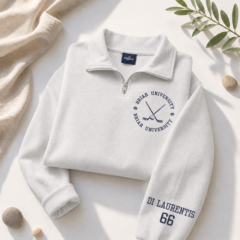 May include: A light gray sweatshirt with a quarter-zip collar. The sweatshirt features a navy blue circular emblem with crossed hockey sticks and the words "Briar University." The sleeve displays "Di Laurentis 66" in navy blue. The sweatshirt is folded on a white surface.