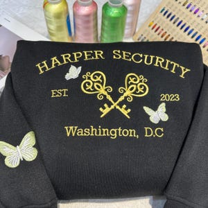 May include: Black sweatshirt with gold embroidery of the text "HARPER SECURITY", "EST. 2023", "Washington, D.C.", two keys crossed, and two butterflies.