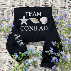 May include: Black cropped sweatshirt with white embroidered text and seashell designs. The text reads "TEAM CONRAD" and "The Summer I Turned Pretty". Features a starfish, a seashell, and a scallop shell.