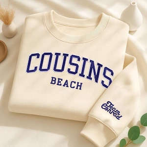 Peut inclure: Sweat-shirt crème avec l'inscription "COUSINS BEACH" en bleu marine sur la poitrine. La manche porte l'inscription "Team Conrad" en bleu marine. Le sweat-shirt a un col rond et des poignets côtelés.