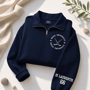 May include: Navy blue sweatshirt with a quarter-zip collar. The front features a white Briar University logo with crossed hockey sticks. The sleeve displays "DI LAURENTIS 66" in white lettering. The sweatshirt is folded on a light-colored surface.