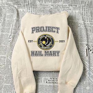 Op de afbeelding: Crèmekleurige sweatshirt met de tekst "PROJECT HAIL MARY" in zwarte en gouden letters. Een cirkelvormig embleem met de woorden "SAVE HUMAN EARTH" is gecentreerd onder het woord "PROJECT". De sweatshirt heeft een ronde hals en lange mouwen.