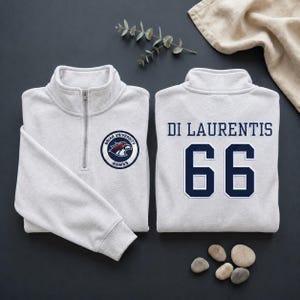 Op de afbeelding: Twee lichtgrijze sweatshirts met een kwart rits. Eén heeft het Briar University Hawks-logo. De andere heeft "DI LAURENTIS" en het nummer "66" in marineblauw. Gepresenteerd op een donkergrijze ondergrond.