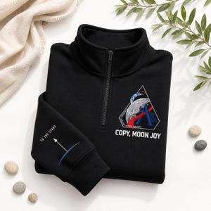 Pode incluir: Camisola preta com fecho de correr a um quarto e gola alta. A camisola apresenta um patch bordado colorido com um design de lua e foguete, e o texto "COPY, MOON JOY". A manga tem o texto "TO THE STARS" e um gráfico.