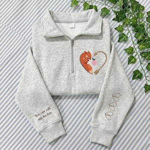 May include: A light gray heathered quarter-zip sweatshirt featuring an embroidered orange fox curled into a heart shape with an arrow and a small pink apple. The text "You can call me Archer." is on the right sleeve with three small heart outlines.