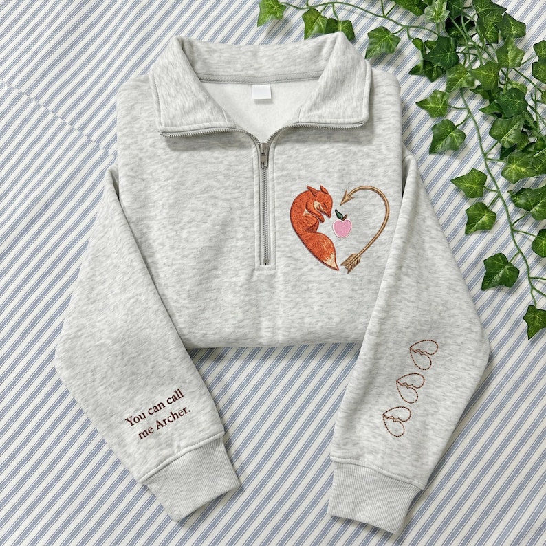 May include: A light gray heathered quarter-zip sweatshirt featuring an embroidered orange fox curled into a heart shape with an arrow and a small pink apple. The text "You can call me Archer." is on the right sleeve with three small heart outlines.