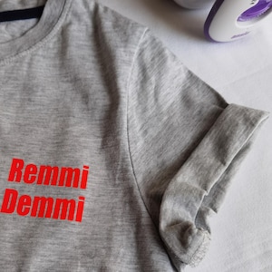 Könnte beinhalten: Ein graues T-Shirt mit dem Aufdruck "Remmi Demmi" in Rot auf der Vorderseite.