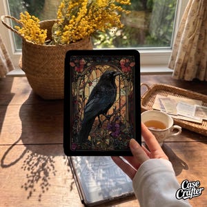 May include: A black e-reader case featuring a vibrant stained glass design of a crow perched on a branch. The artwork includes intricate floral patterns in shades of red, orange, and purple, framed by an arched border against a dark background.