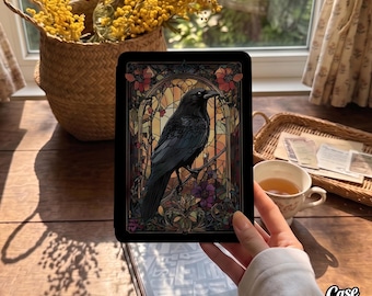 May include: A black e-reader case featuring a vibrant stained glass design of a crow perched on a branch. The artwork includes intricate floral patterns in shades of red, orange, and purple, framed by an arched border against a dark background.