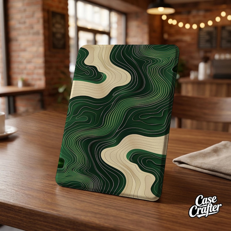May include: A green and cream topographic design adorns a rectangular product, possibly a tablet case. The design features layered lines in varying shades of green and cream, creating a textured, abstract pattern. The product is displayed on a wooden table.