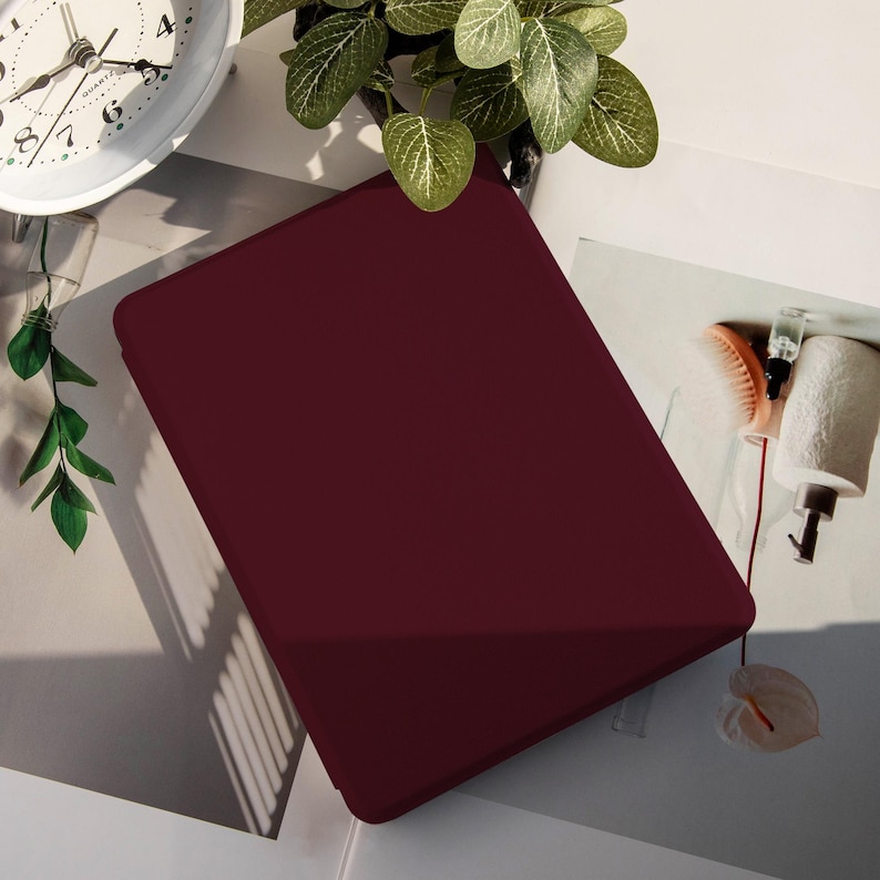 May include: A burgundy tablet case. The case is rectangular and has a smooth, matte finish. The image also shows a white clock, green leaves, and other decorative items.